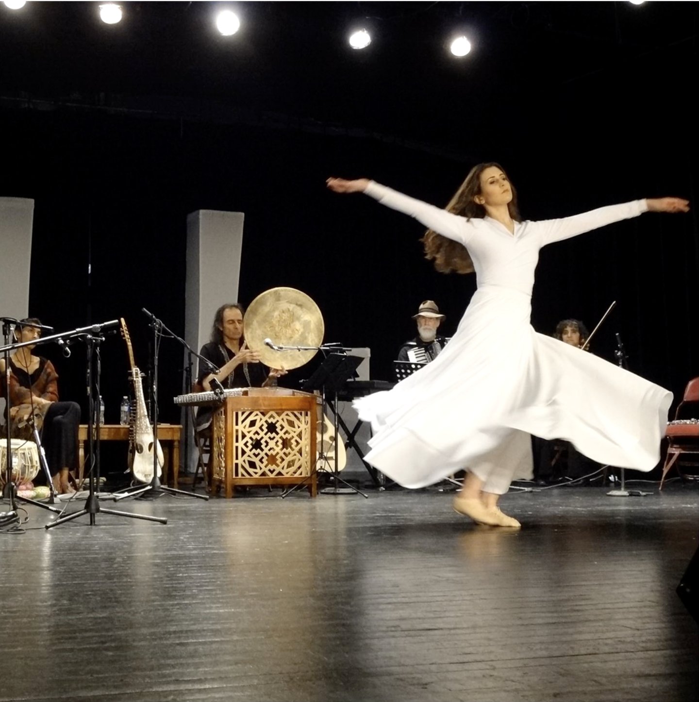A whirling dancer in white spins before the orchestra on a concert stage, with daf, tabla, accordion and violin behind her